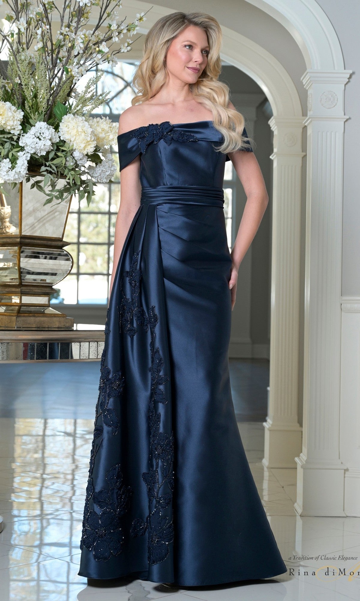 A woman in a navy Long Formal Dress (Rina Di Montella RD3504) with an off-the-shoulder neckline, ruched waistband, and floral embroidery stands indoors near a flower arrangement and white columns.